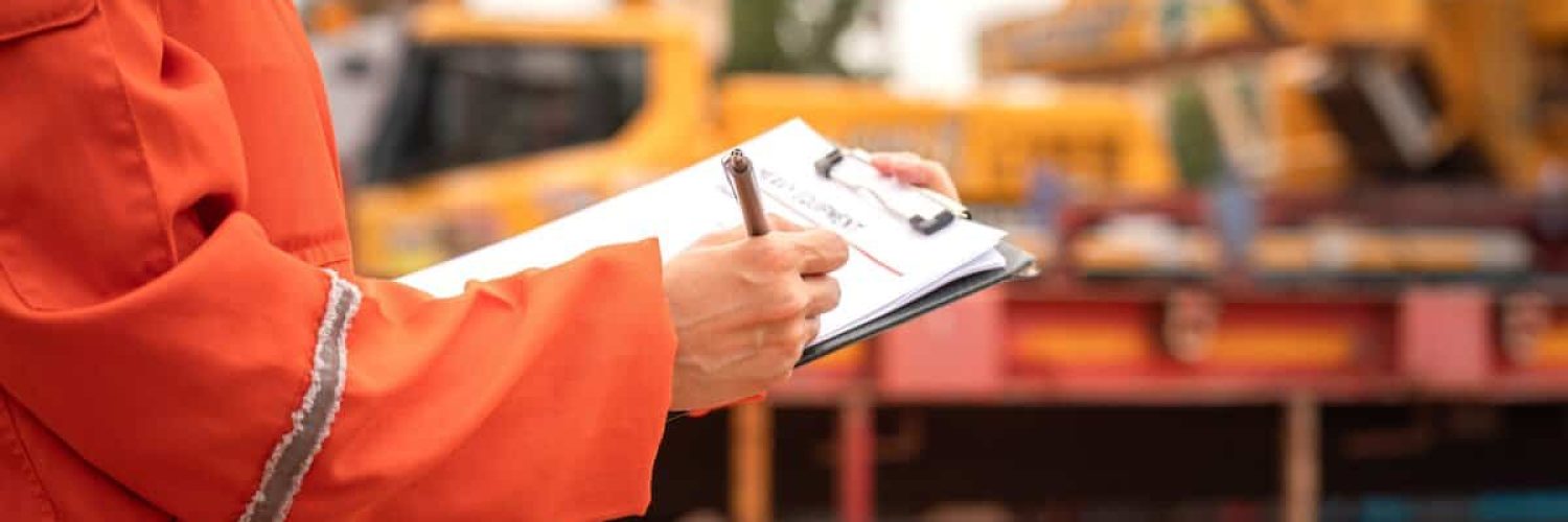 Close-up at the operation supervisor is checking on heavy machine and equipment checklist form with crane truck as blurred background. Industrial safety working in action, selective focus.